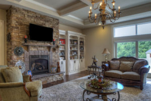 An open living room with a custom built stone fireplace, a chandelier, built in shelving, with a couch and chair. There is a window looking out to the backyard.