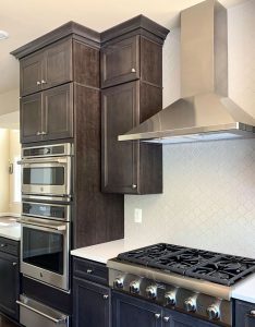 A custom built kitchen a white backsplash, dark brown cabinets, a double oven, and luxury gas stove.