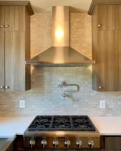 A kitchen with luxury gas stove, cooktop vent, white tile backsplash, and beautiful light brown-grey cabinets.
