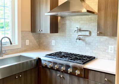 A kitchen with luxury gas stove, cooktop vent, white tile backsplash, stainless steel sink, and beautiful light brown-grey cabinets.
