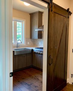 A dark brown barn door, halfway open showing the kitchen with a stainless steel sink and light grey-brown cabinets.