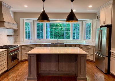 A custom built kitchen with a large oversized island counter, sleek black metal ceiling lamps, white granite counters, and a large window above the sink.