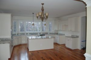 An open, white kitchen with grey granite counters. There is brown hardwood flooring and a medium sized chandelier.