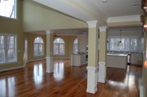 An unfurnished, but finished open living room, kitchen, and dining area. There are a few pillars . Windows surround the interior walls with views of a forest.