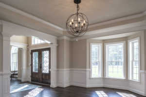 Living room area with light fixture, dark brown floors and front door. A large window brings in natural light.