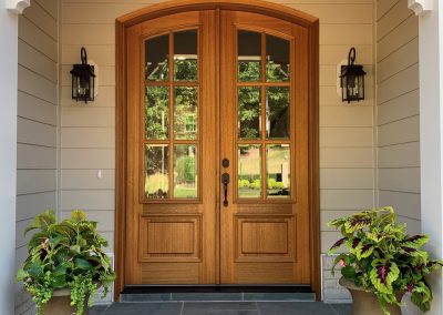 Medium to light brown front door with black glass sconces and plants to the right and left sides.